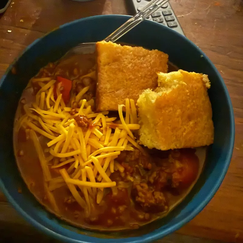 A bowl of chili topped with shredded cheese, with cornbread.